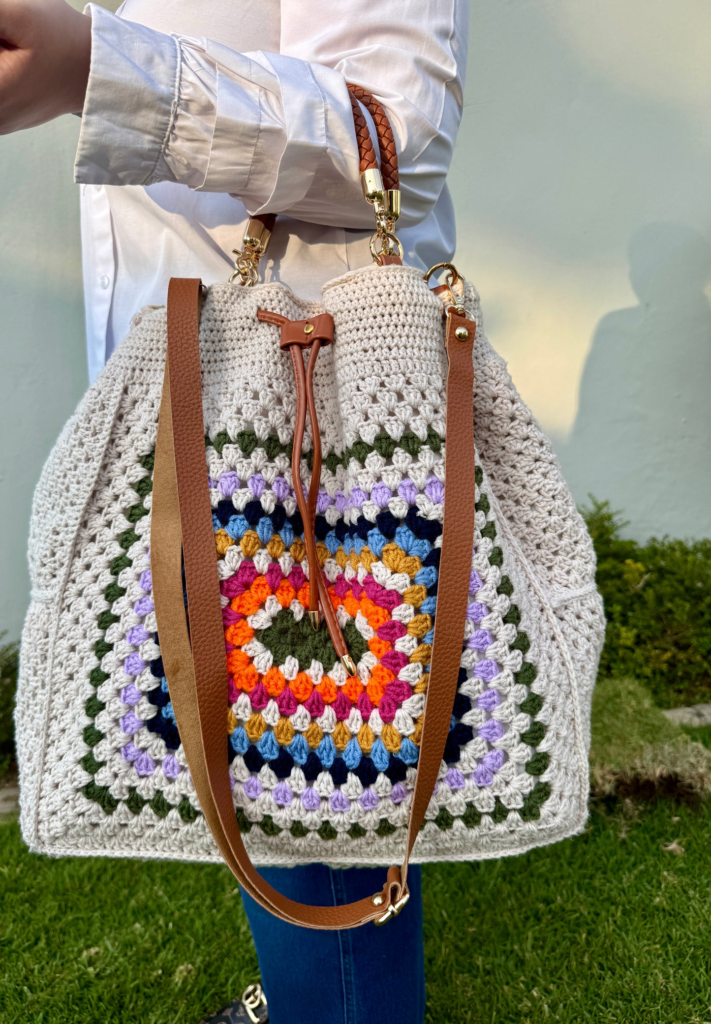 Crocheted bag with colorful pattern and brown straps held by a person outdoors.