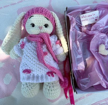 Pink crochet bunny doll with hat and scarf, sitting beside a pink tissue-wrapped gift box.
