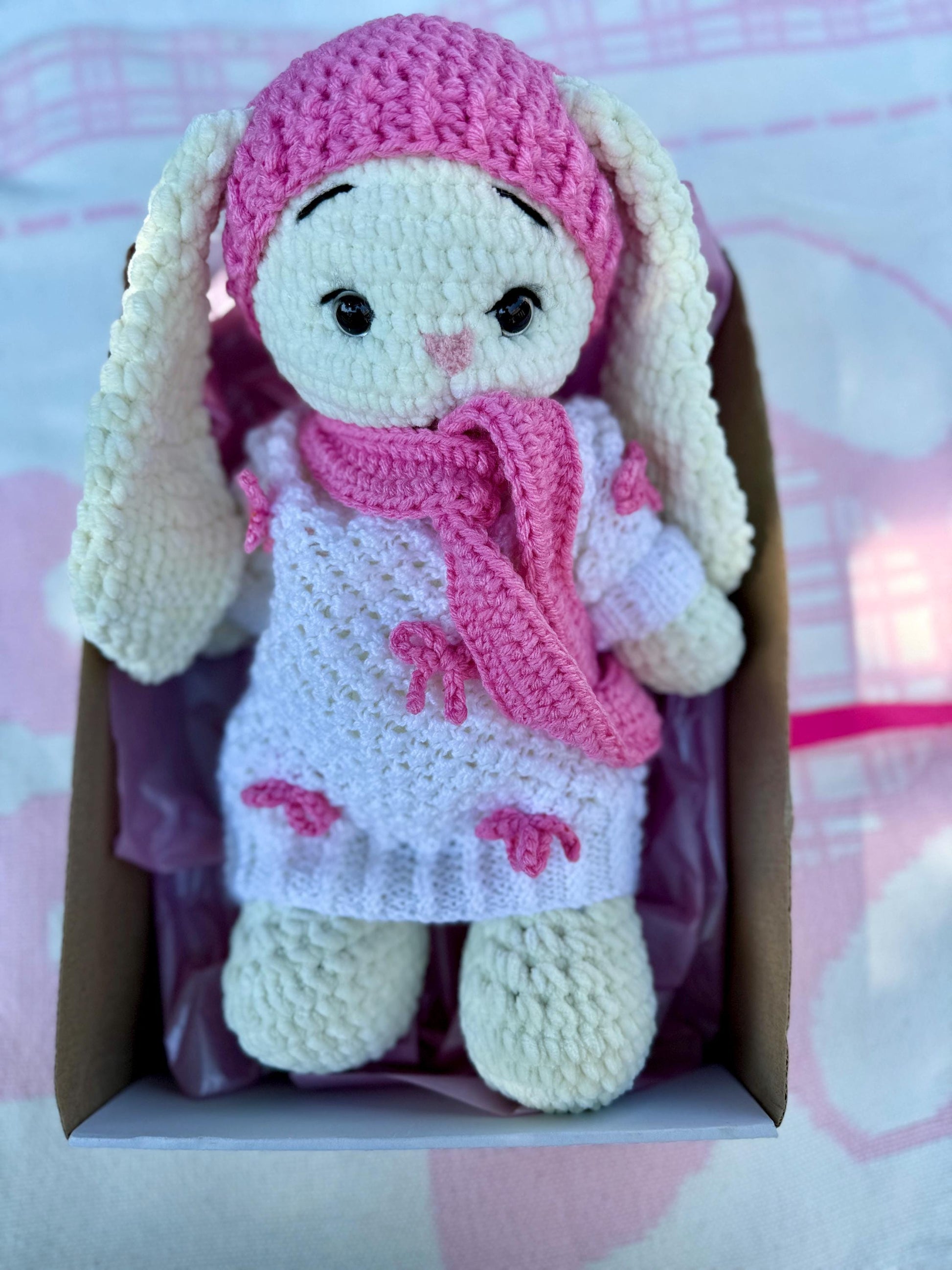 Pink crochet bunny doll in white dress with small bows, hat, and scarf inside a gift box.
