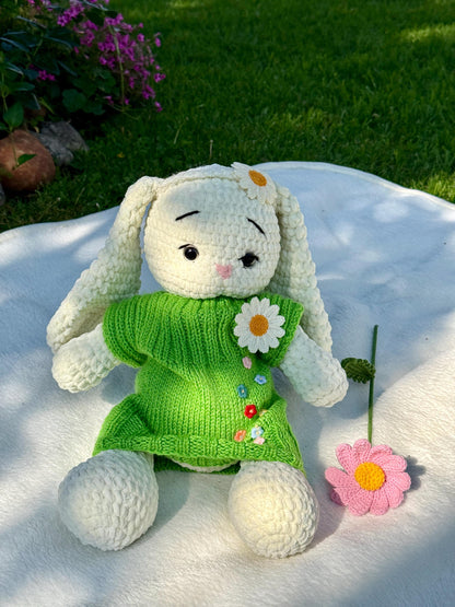 Crochet bunny doll wearing a green knitted daisy dress sitting on a white blanket in sunlight