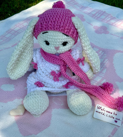 Soft crochet bunny with pink hat and matching scarf wearing a white dress decorated with bows.
